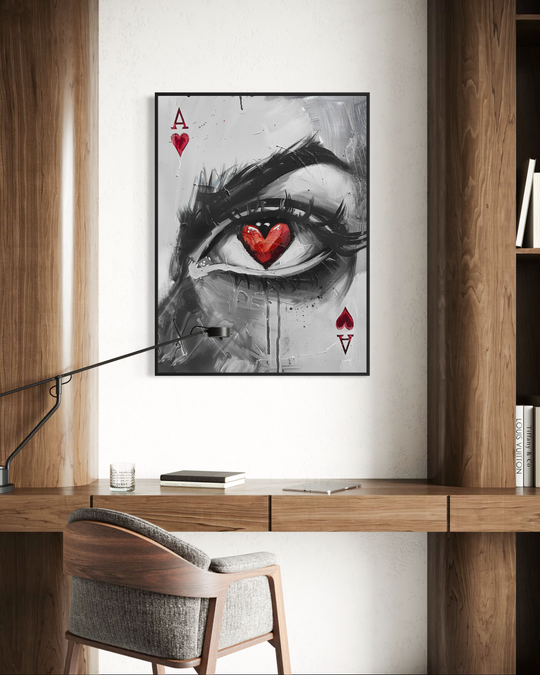 A framed black and white portrait featuring a single detailed eye with a red heart-shaped pupil, styled like an Ace of Hearts playing card, hanging on a clean minimalist wall above a wooden desk setup with a chair, books, and a glass. The artwork combines expressive brushstrokes with bold red accents, creating a striking focal point in a warm contemporary interior.
