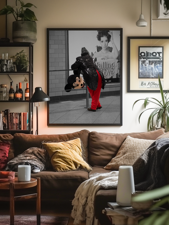 A cozy, eclectic living room with a brown sectional sofa, layered pillows and blankets, and warm ambient lighting. The large framed poster above the couch features a striking street-style photo of a performer resting in costume, adding humor and character to the inviting space. Surrounding elements like books, plants, and decor make the room feel personal and lived-in.