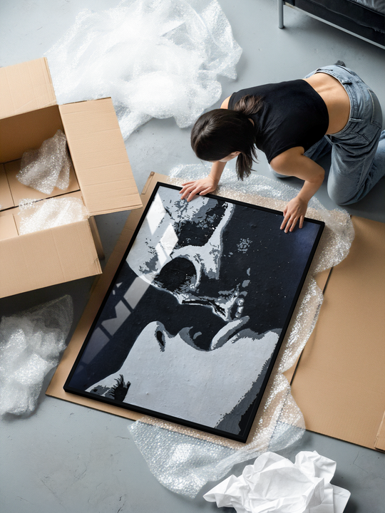 A woman kneels on the floor unboxing a large framed black and white street art print. The artwork shows a close up portrait of a woman licking a skull, rendered in a gritty stencil banksy-inspired style. The frame rests on bubble wrap and cardboard packaging, surrounded by unpacked materials.
