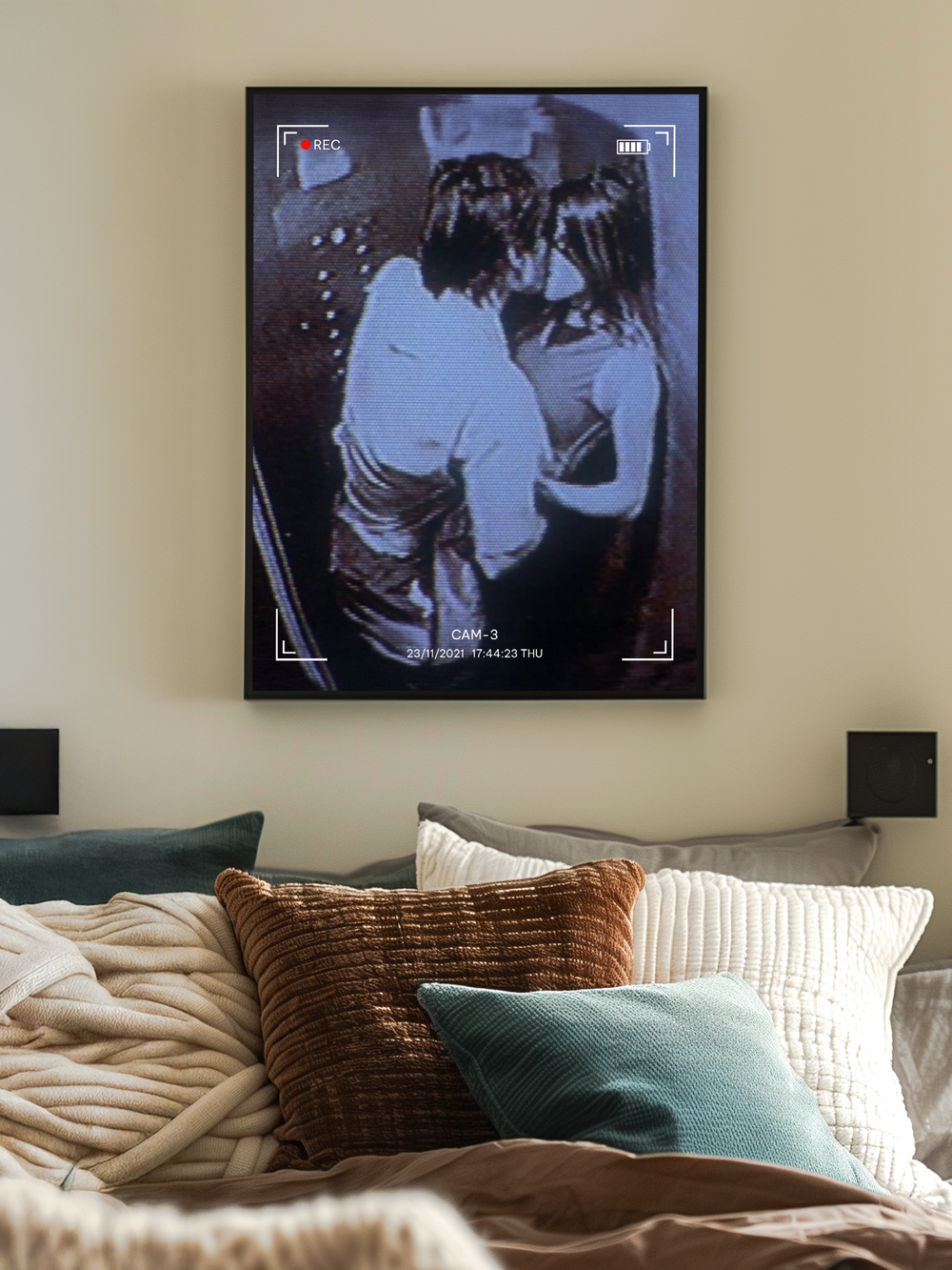 A vertical poster showing a grainy CCTV-style image of a couple kissing, complete with a REC symbol and timestamp, hangs centered above a cozy bed. The bed is styled with layered pillows in warm earth tones, soft blankets, and natural light adding warmth to the neutral-colored room. The intimate setup creates a raw and emotional atmosphere appealing to a young adult audience.