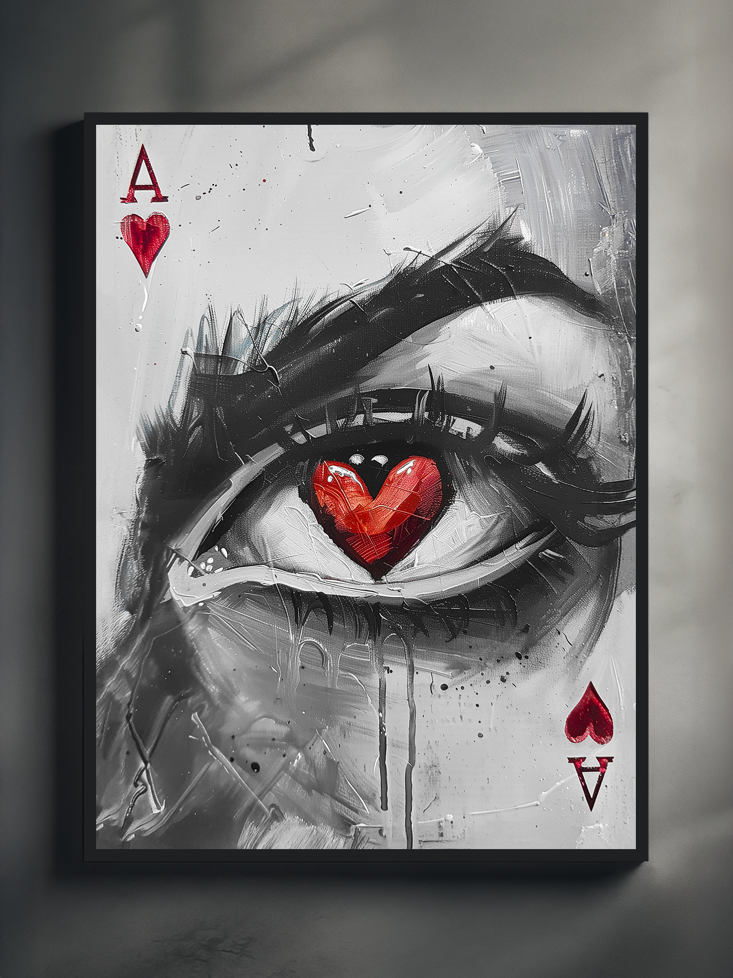 A modern wall-mounted poster showing a surreal black-and-white eye with a red heart-shaped pupil, designed in the style of an Ace of Hearts playing card. The artwork is framed in black and photographed with angled lighting, creating shadows and a minimalist gallery-like setting.