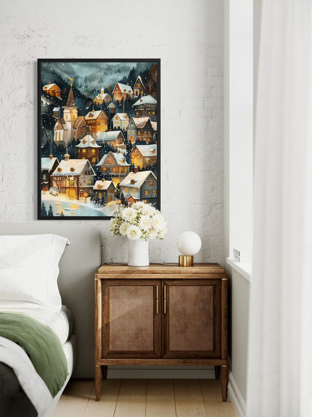 A winter village wall art print hangs on a textured light wall above a small bedroom cabinet. The artwork shows a snowy mountain village with warm glowing windows, detailed houses and falling snow. Soft bedding, a vase of white flowers and gentle natural light create a peaceful and cozy atmosphere around the artwork.
