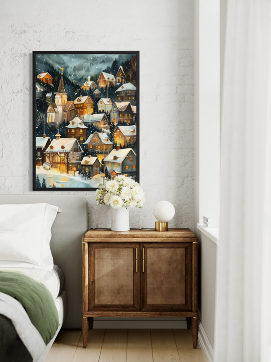 A winter village wall art print hangs on a textured light wall above a small bedroom cabinet. The artwork shows a snowy mountain village with warm glowing windows, detailed houses and falling snow. Soft bedding, a vase of white flowers and gentle natural light create a peaceful and cozy atmosphere around the artwork.
