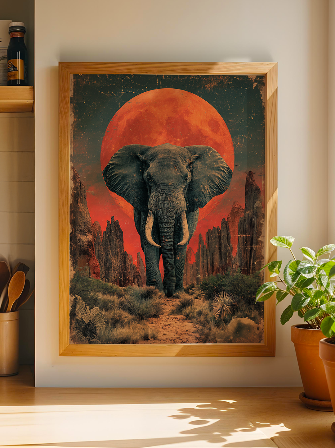 Framed artwork featuring a majestic elephant walking through a desert path surrounded by cacti and rock formations, with a dramatic red full moon rising in the background, blending wildlife and surreal fantasy elements.
