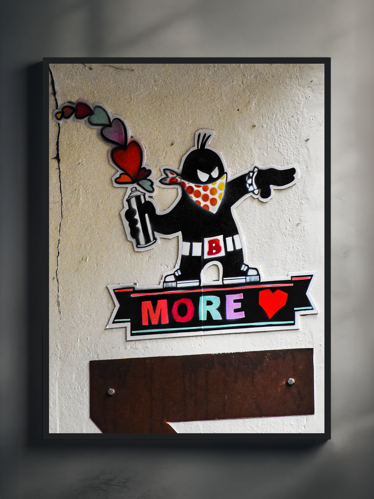High-quality wall art print featuring a photographed Berlin street art piece with a cartoon-style graffiti character spraying colorful hearts. The artwork includes bold stencil details and the message “MORE LOVE” on a weathered concrete wall, framed in black for a modern urban look. Perfect for fans of graffiti art, pop culture decor, and contemporary wall prints with a street vibe.