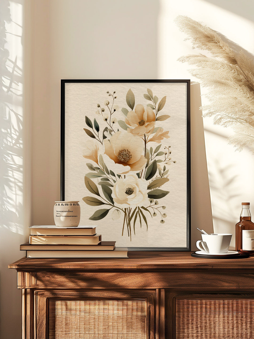 A vertically framed poster with a floral illustration stands centered on a wooden cabinet, surrounded by books, a white cup, and dried pampas grass. The room is warmly lit by natural sunlight, creating soft shadows and a cozy, elegant atmosphere.