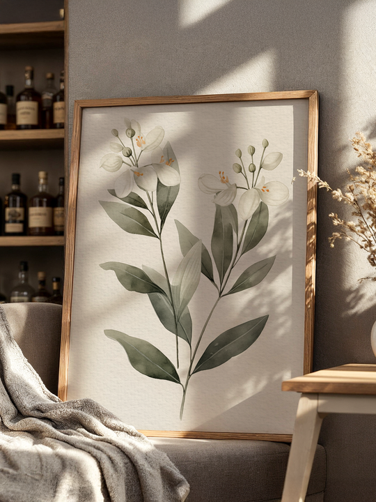 A serene botanical art print showcasing delicate white flowers and large green leaves in a light wooden frame. The artwork leans casually on a couch in a softly lit corner, surrounded by cozy textures, neutral tones, and a wooden shelf with bottles and books, creating a peaceful and stylish interior scene.