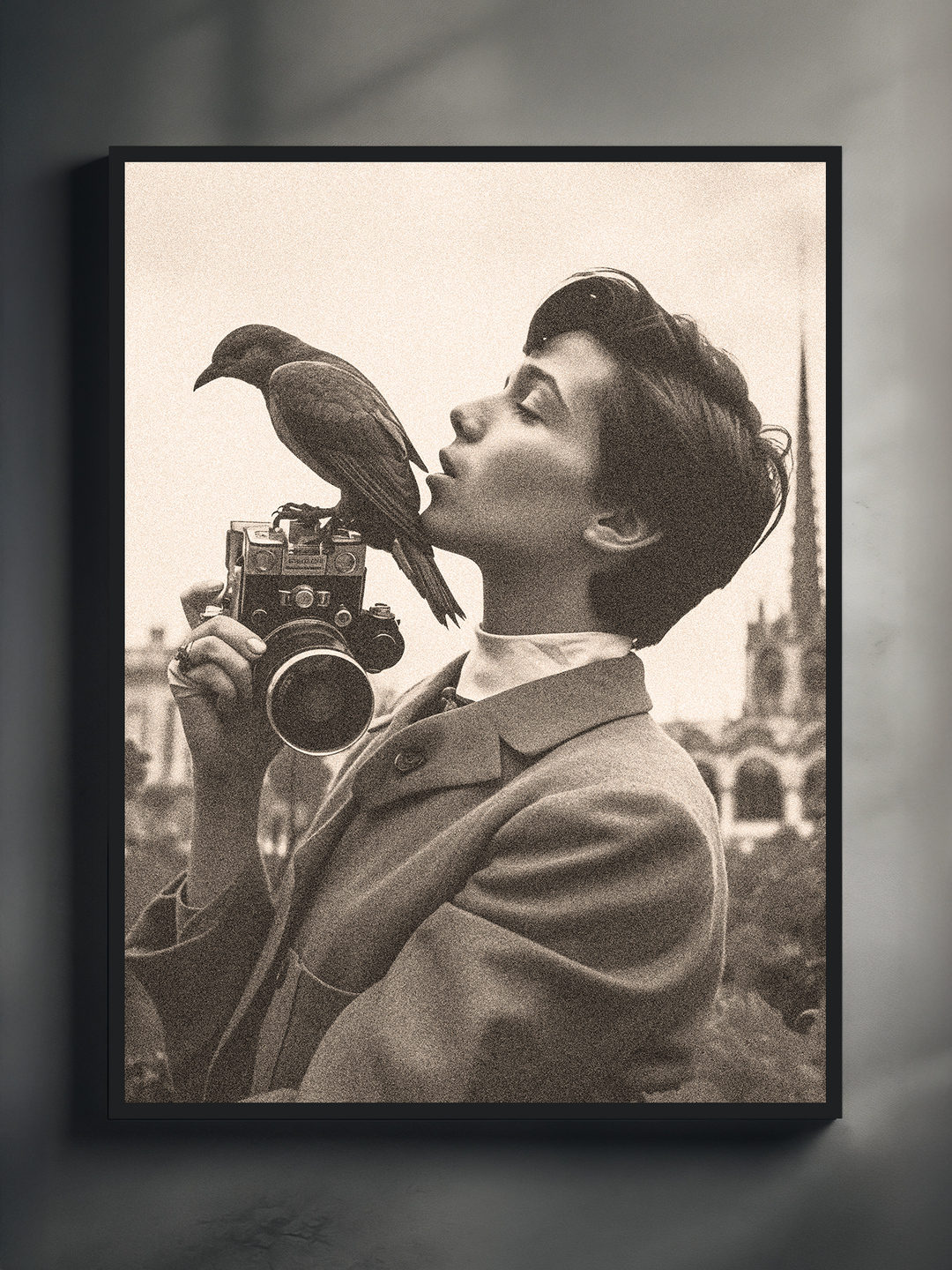 A black-and-white portrait of a young man holding an old-fashioned camera, while a bird is perched gracefully on top of it. The man leans in as if to kiss the bird, creating a poetic and serene moment. He wears a vintage suit, and the blurred background features Gothic architecture, adding a timeless European flair to the composition. The artwork is printed on textured material and framed with a dark border.