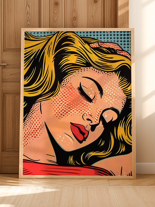 A colorful pop art poster featuring a close-up of a blonde woman with red lips and closed eyes, displayed in a light wooden frame, leaning against a wooden wall on a bright wooden floor with sunlight casting soft shadows.