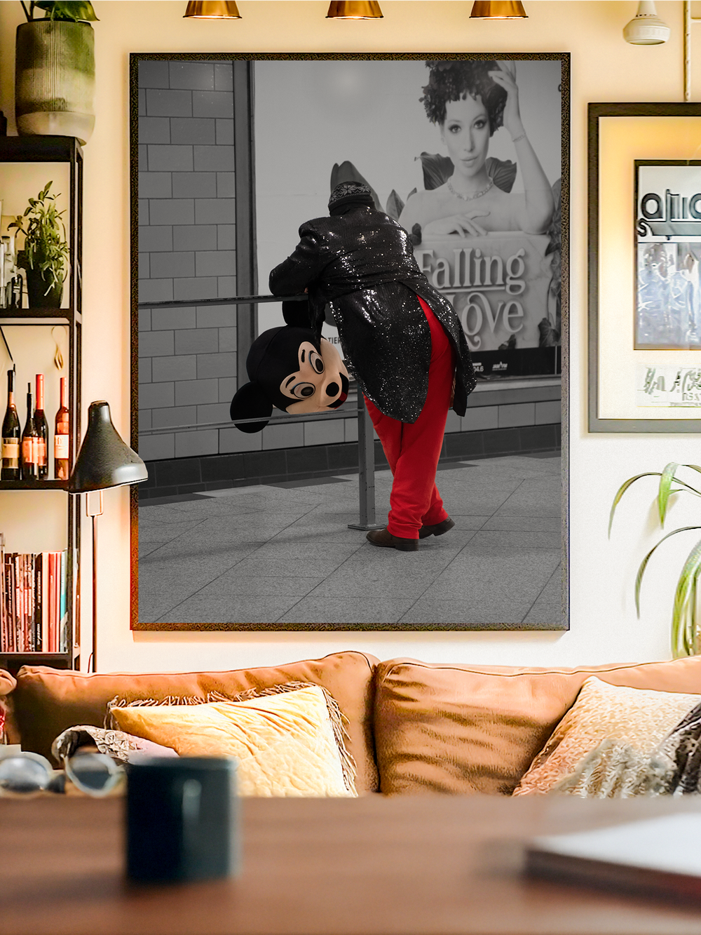 A framed artwork showing a performer in a sequined black jacket and red pants, holding a Mickey Mouse head while leaning against a pole in front of a grayscale billboard that reads “Falling in Love”. The bold contrast between color and monochrome creates a striking pop art effect. The poster hangs in a modern living room with a tan sofa, books, plants, and warm ambient lighting, blending edgy urban art with cozy interior design.