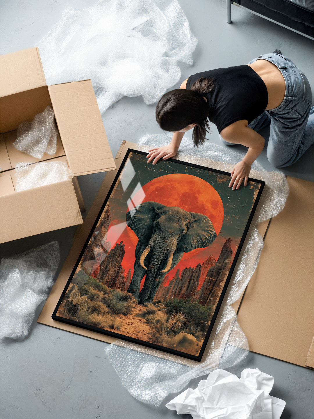 A woman kneels on the floor while unboxing a large framed poster surrounded by bubble wrap and cardboard. The artwork features a powerful elephant walking forward through a rugged desert landscape with dry plants and sharp rock formations. Behind the elephant rises an enormous red moon, creating a surreal, dramatic atmosphere. Warm orange-red tones blend with deep greens and earthy shades, giving the scene a mix of wildlife energy and fantasy aesthetics. The print is framed in a sleek black frame with gloss