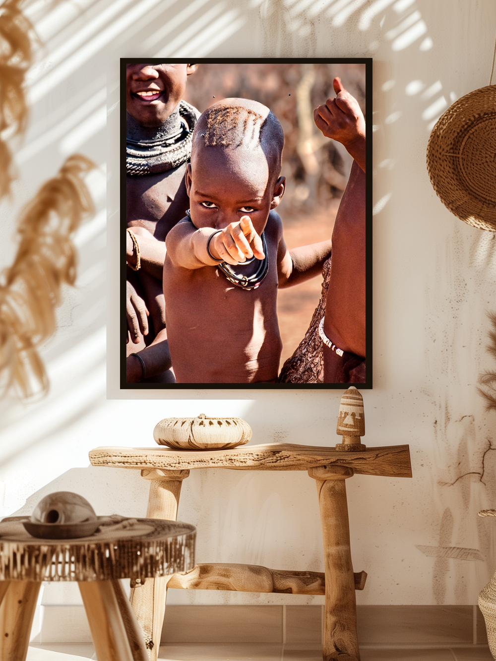 A powerful framed poster showing a young child with a traditional tribal hairstyle and jewelry is displayed on a white wall in a warm, ethnic-style interior. The room features handcrafted wooden furniture, woven decorations, and natural textures, creating a rustic and authentic atmosphere that highlights indigenous culture.