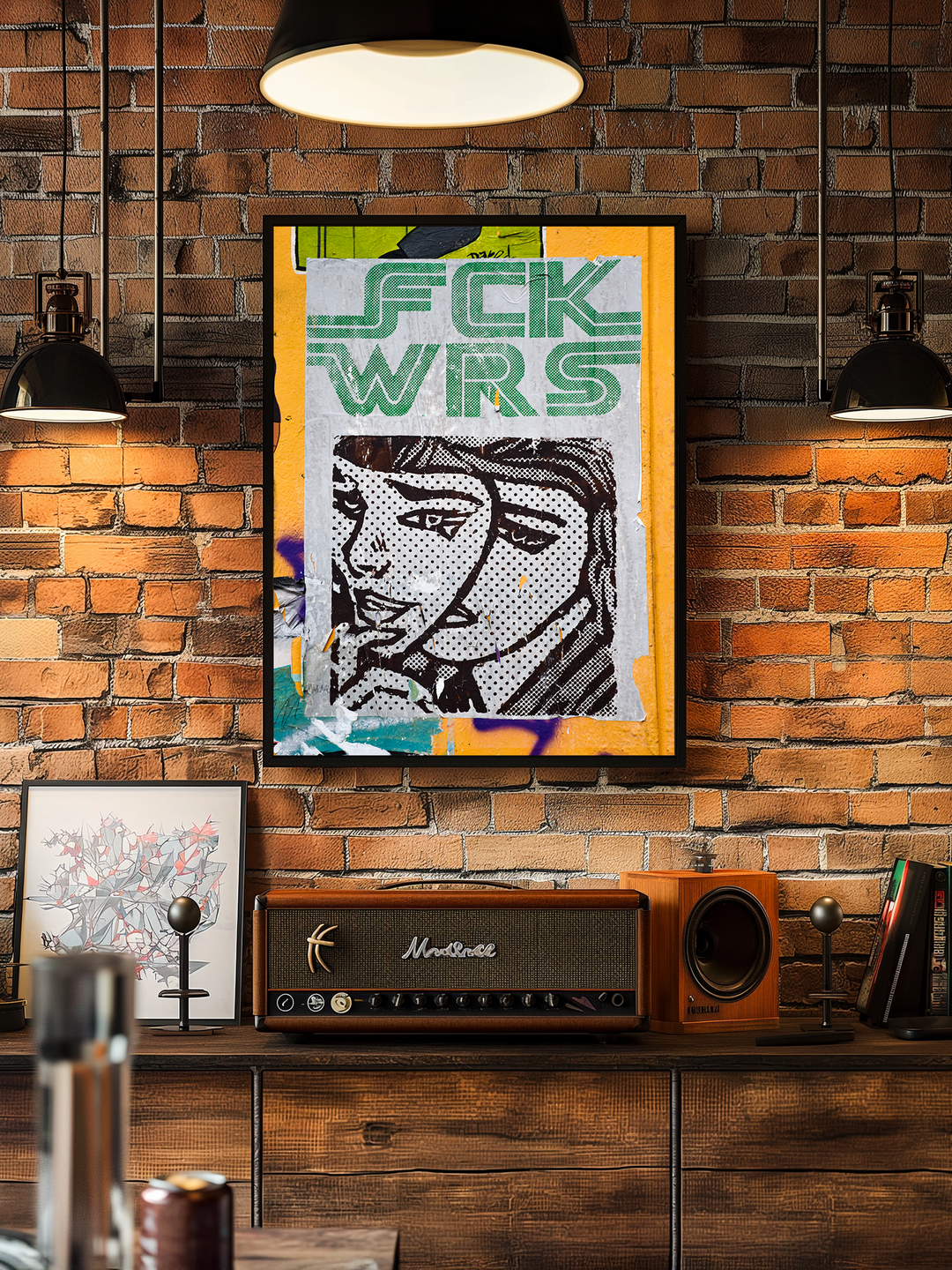 Framed pop art poster featuring bold graffiti-style lettering and a comic-style female face, hanging on an exposed brick wall in a stylish loft with industrial lighting, wooden sideboard, retro amplifier, and bookshelf.