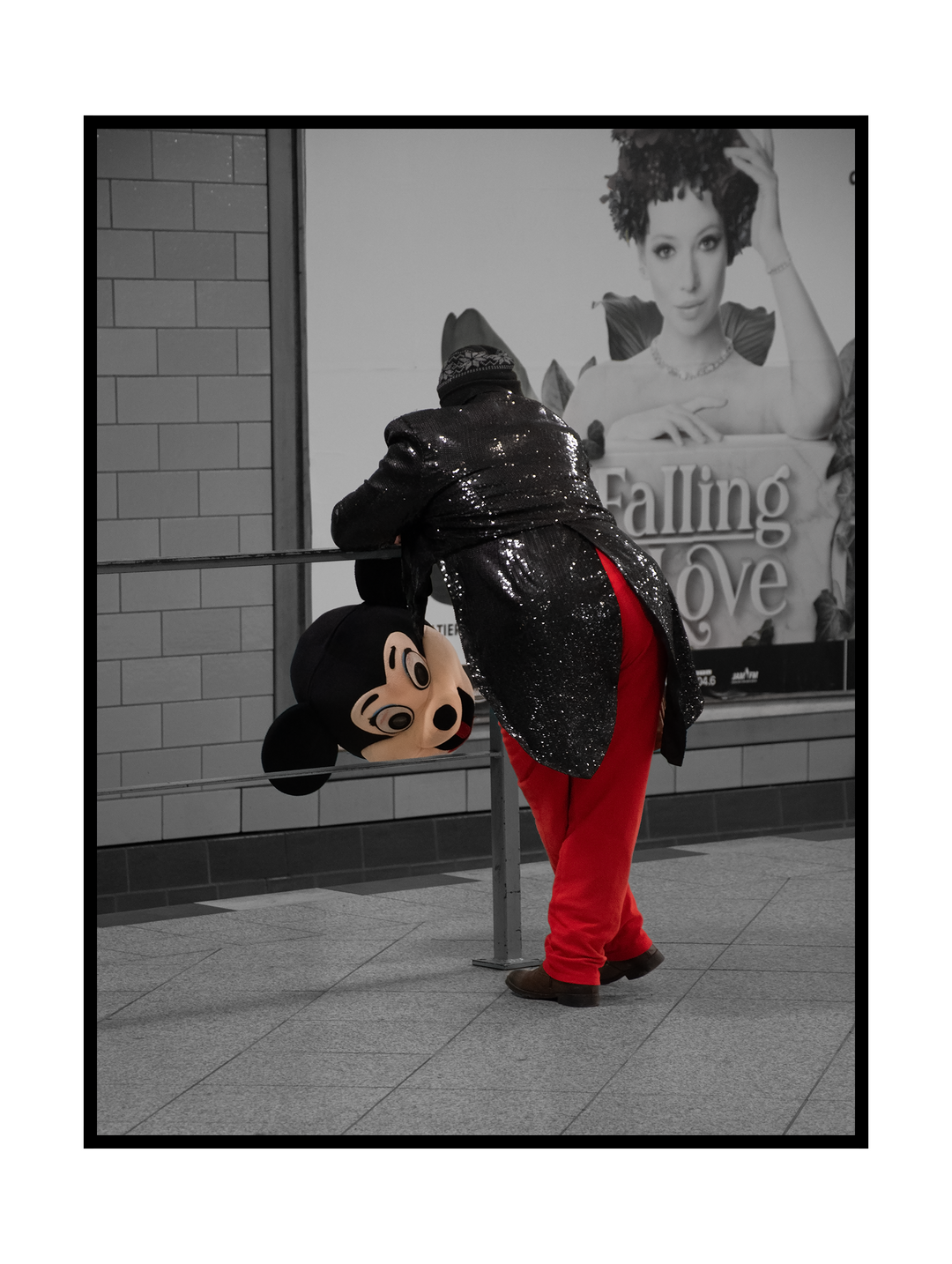 Striking urban wall art poster featuring a surreal street scene of a person in a Mickey-style costume leaning over a railing, holding the character head, set against a grayscale city backdrop. Selective color highlights the bright red pants, creating strong contrast and visual tension. Conceptual street photography artwork designed as modern wall art for contemporary interiors, studios or urban-inspired spaces.