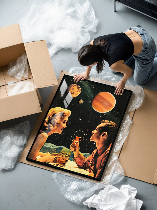 A woman kneels on the floor unpacking a framed art print. The poster shows a retro-futuristic couple sharing a drink at a table while large planets float in a starry sky behind them. The colors and vintage styling give it a classic retro romance feel. The framed print lies on packaging material next to an open cardboard box.
